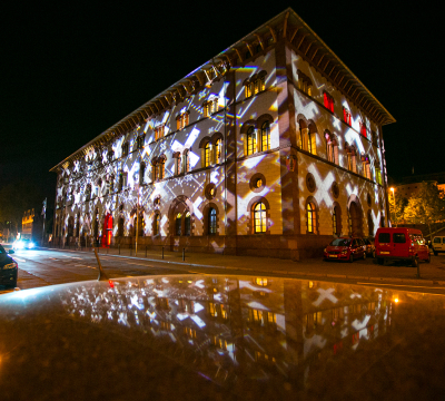Die Fruchthalle vom Schillerplatz aus. Es ist Nacht und sie wird mit "X'en" bestrahlt.Die Lichter spiegeln sich auf dem Autodach hinterdemk Der Fotograf steht.