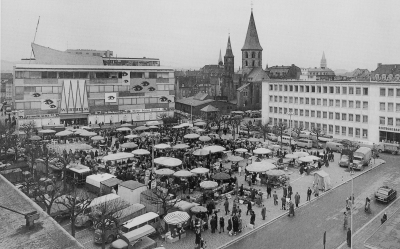 Viele Menschen besuchen den Wochenmarkt auf dem Stiftsplatz. Die offenen Sonnenschirme wirken wie ein Dach aus hellen Tupfern und dazwischen erkunden Besucher die Stände. Im Hintergrund ist die Stiftskirche gut zu erkennen.