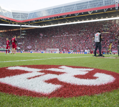 Aufnahme im Inneren des Stadions. Man sieht im Vordergrund das FCK-Logo auf dem Rasen, im Anschluss das Spielfeld mit Spielern des 1. FCK und im Hintergrund die Westtribüne mit Zuschauern.