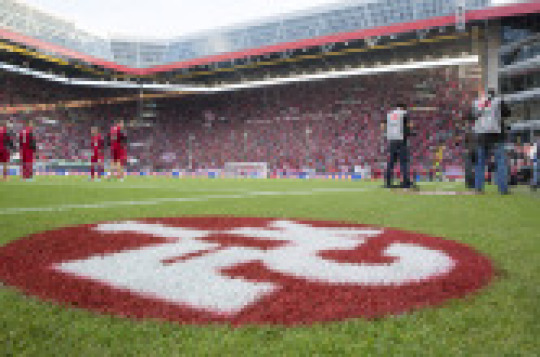 Aufnahme im Inneren des Stadions. Man sieht im Vordergrund das FCK-Logo auf dem Rasen, im Anschluss das Spielfeld mit Spielern des 1. FCK und im Hintergrund die Westtribüne mit Zuschauern. © view - die agentur