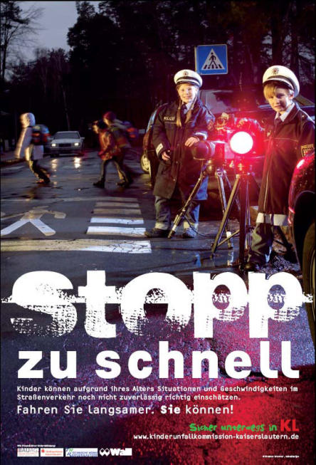 Gezeigt wird ein Plakat mit  großer Schrift - Stopp zu schnell. Zu sehen sind Kinder beim Überqueren eines Zebrastreifens im Hintergrund. Davor stehen zwei Kinder in Polizeiuniform , die eine Radarfalle bedienen.