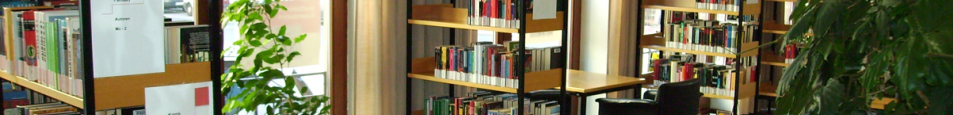 Zwischen den Bücherregalen stehen große, einladende, begrünte Blumenkästen und Vasen. Ein gemütlicher Sessel an einem Tisch lädt zum Verweilen und Schmökern ein