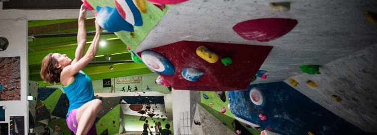 junge Frau hängt frei an Boulderwand - zwei junge Männer, auf dem Boden sitzend, schauen zu