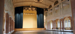 Durch die halbrunden Fenster dringt Licht von außen in den großen Saal der Fruchthalle. Ein schwerer Vorhang ist halb geöffnet und gibt den Mittelteil der Bühne frei. Obwohl der Saal leer ist wirkt er durch den balkonartigen Aufbau und die schlichten, marmorierten Säulen sehr festlich.
