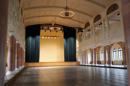 Durch die halbrunden Fenster dringt Licht von außen in den großen Saal der Fruchthalle. Ein schwerer Vorhang ist halb geöffnet und gibt den Mittelteil der Bühne frei. Obwohl der Saal leer ist wirkt er durch den balkonartigen Aufbau und die schlichten, marmorierten Säulen sehr festlich. © Stadt Kaiserslautern
