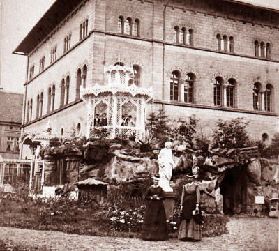 Die Außenansicht der Fruchthalle mit einer Parkanlage und einem mehrstöckigen Pavillon. Zwei Frauen mit langen Kleidern und hüten posieren für das Foto.