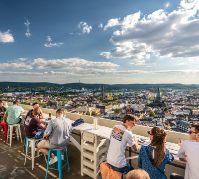 Menschen sitzen auf der Dachterrasse der Sommerlounge des Twenty One. Im Hintergrund sieht man Kaiserslautern.