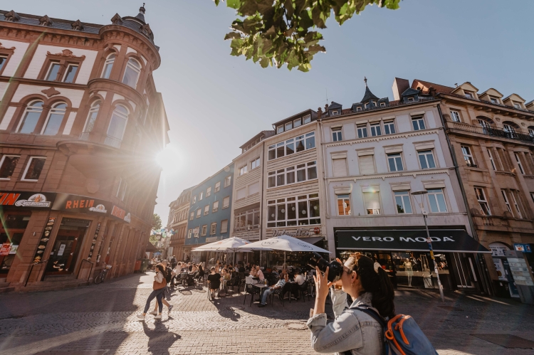 Man sieht den Platz vor der Stiftskirche im Sommer. Im Vordergrund eine Frau, die ein Gebäude fotografiert. Im Hintergrund Außengastronomie. Thomas Linkel, Lizenz CC-BY