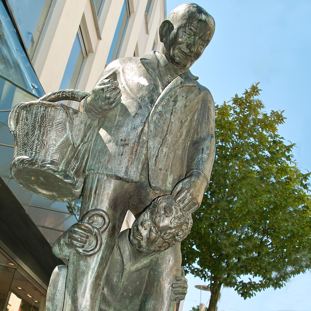 Ein kleiner Junge mit einer Brezel in der Hand schaut durch die Beine des Brezeladam - Bronzefigur  Stadt Kaiserslautern, Lizenz CC-BY